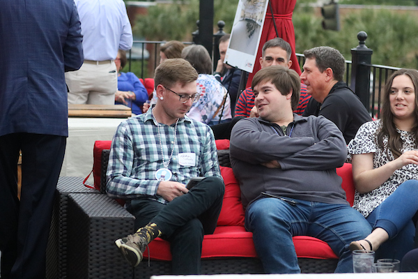 Man with folded arms talking to another ma in plaid shirt sitting on outdoor patio.