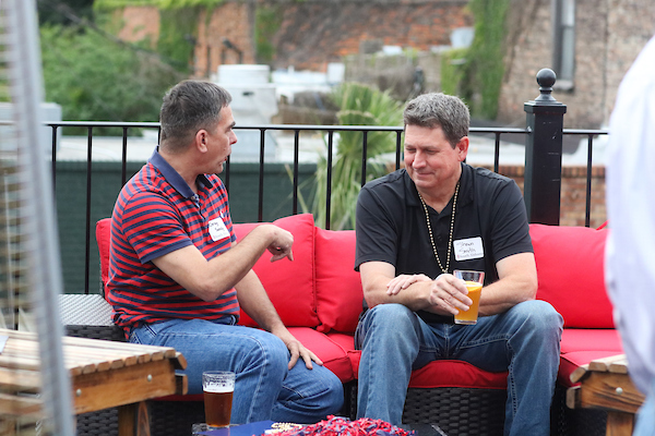 Man in striped shirt talking to man in black shirt sitting on outdoor patio.