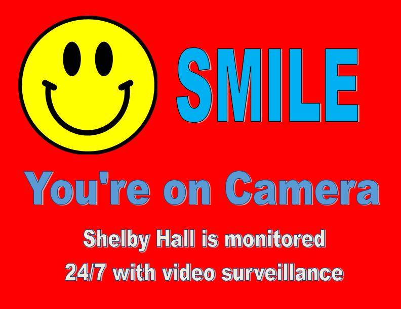 A big smiley face next to the word SMILE in large font. Underneath are the words "You're on Camera" Underneath it says "Shelby Hall is monitored 24/7 with video surveillance