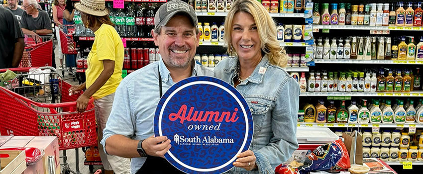 Play House Spices at the grocery store holding up Alumni owned sign.
