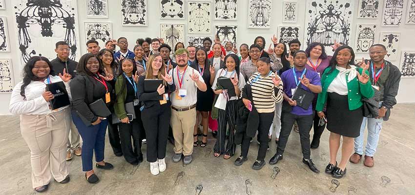 Group of students holding up J hand in front of wall of art.