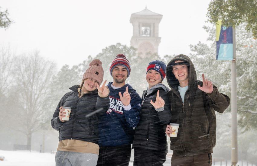 Four students holding up J hand while snow is falling and Moulton tower in background.