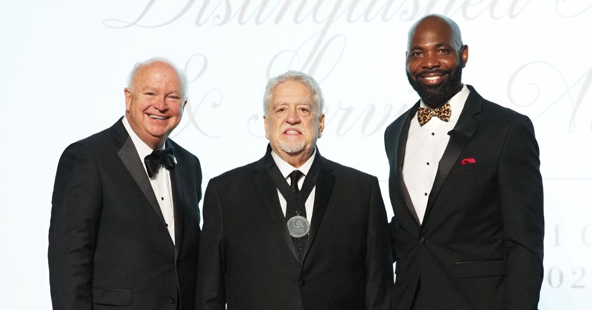 Three men stand on a stage. The center man wears a medal he received in recognition of an award.